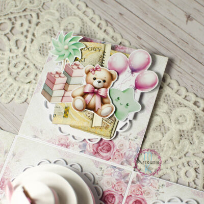 jedna ze ścianek pudełka 3D. Na środku jest siedzący miś z rózową kokardą wokół szyi, po lewej jego stronie są różowo zielone klocki, a nad klockami dziecięcy wiatraczej. Po prawej stronie misia jest zielona gwiazdka a nad nią różowe balony. Pod grafikami znajduje się miejsce na włożenie pieniędzy