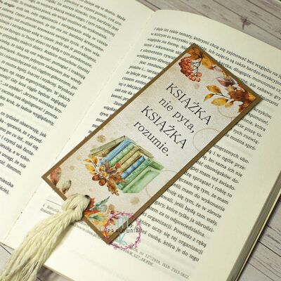 Zakładka prostokątna z białym frędzlem na końcu. Na środku zakładki jest napis Książka nie pyta, książka rozumie, pod tekstem jest grafika książek z jesiennymi liśćmi. U góry jak i na dole zakładki znajduje się grafika jesiennych liści.