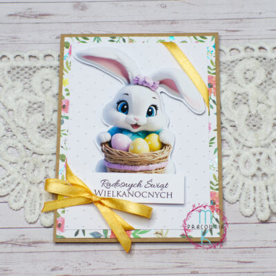 kartka wielkanocna jednokartkowa na środku znajduje się biały zajączek trzymający w łapkach koszyk z pisankami. Całość przewiązana jest pomarańczową wstążką