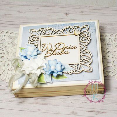 drewniane pudełko na którego wieczku w centralnej części jest ramka i napis w dniu ślubu. W dolnymlewym rogu znajdują się biało niebieskie kwiaty z białą wstązką