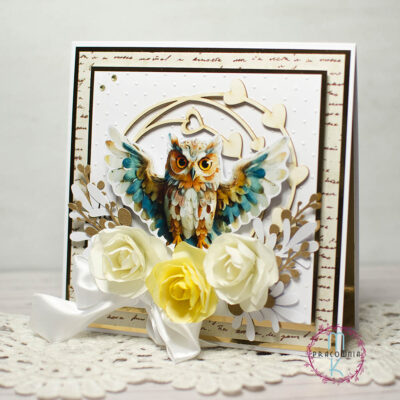 Kartka kwadratowa. W środkowej części znajduje się sowa z rozpostartymi skrzydłami, pod nią kwiaty 3 D w odcieniach kremu i żółtego. Wokół sowy znajduje się ramka z serduszkami