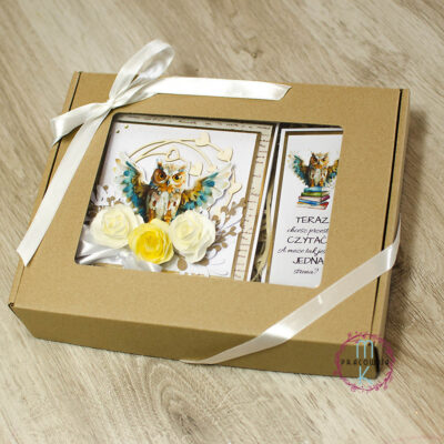 Kartka kwadratowa. W środkowej części znajduje się sowa z rozpostartymi skrzydłami, pod nią kwiaty 3 D w odcieniach kremu i żółtego. Wokół sowy znajduje się ramka z serduszkami. Obok kartki znajduje się zakładka z tą samą sową i z cytatem. Całość zapakowana w brązowe pudełko i przewiązane wstążką