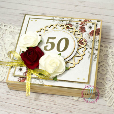 pamiątka na złote gody w drewnianym pudełku. Na środku cyfra 50 w otoczeniu tekturkowej serwetki. Z lewej strony w narożniku kwiaty białe i bordowe ze złotą wstążką. Pamiątka w kolorach złota i czerwieni