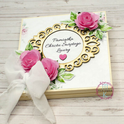 Drewniane pudełko na Chrzes Święty. Na środku ozdobna ramka z tekturki a wniej napis Pamiątka Chrztu Świętego imię dziecka. W prawym górnym rogu umieszczona jest wystająca róża w kolorze różowym. Takie same dwie róże zostały umieszczone w lewym dolnym rogu razem z białą kokardą