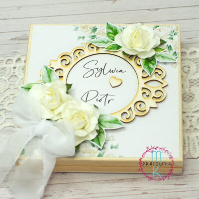 drewniane pudełko, na wieczku którego w centralnym miejscu znajduje się ozdobna ramka z imionami pary młodej. W górnym prawym rogu znajduje się kremowa wystająca róża. W dolnym lewym boku znajdują się dwie kremowe wystające róże i biała szeroka wstążka dopełniająca całości